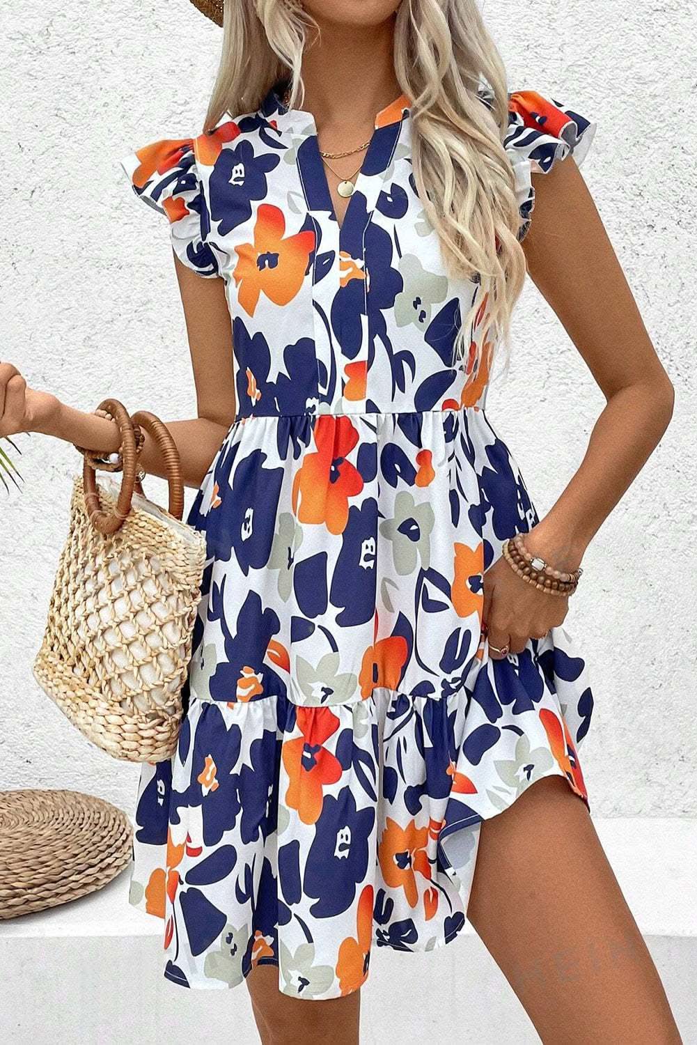 Woman wearing orange floral print notched neck flutter sleeve mini dress with ruffle tiered skirt and holding a woven handbag