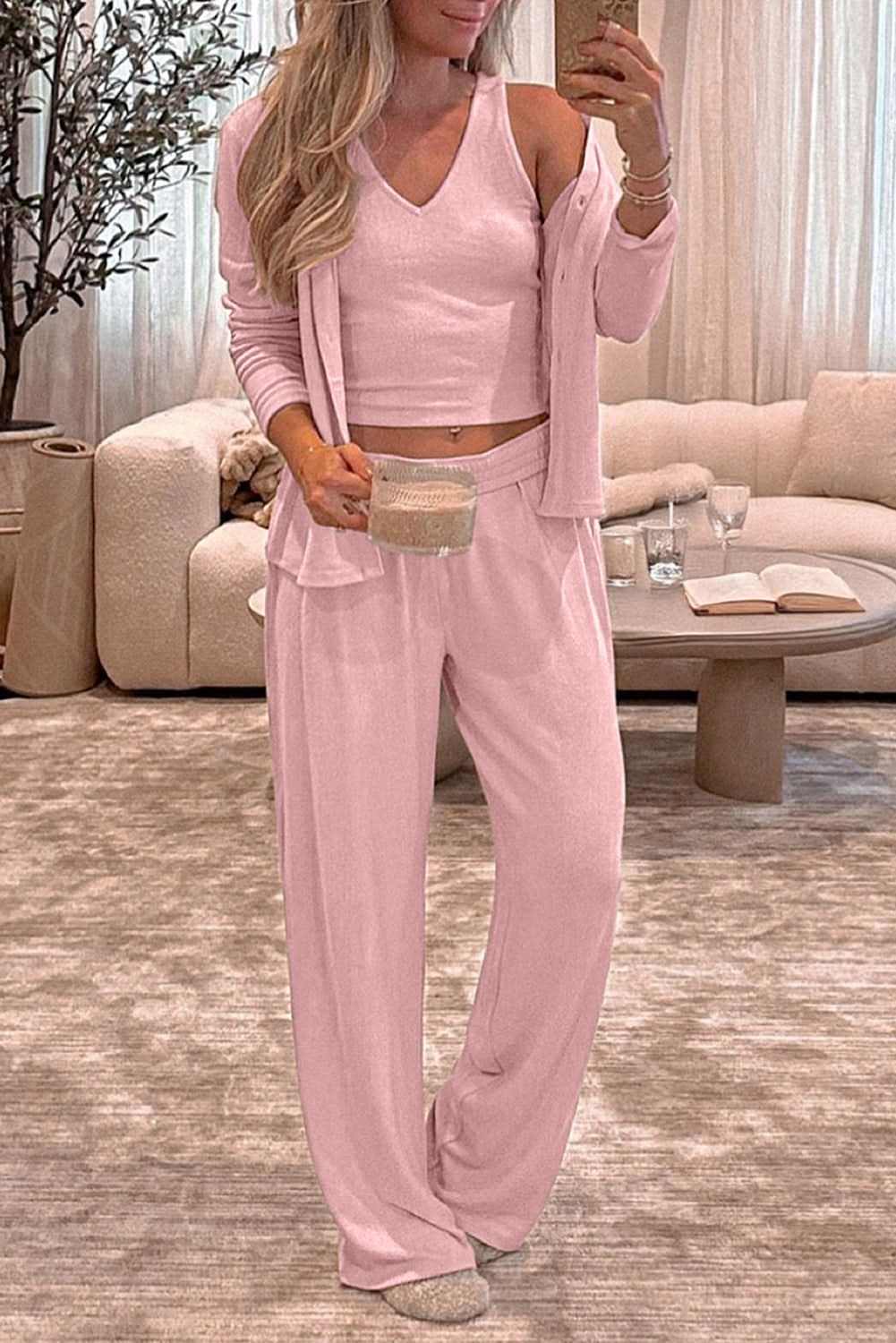 Woman wearing light pink 3-piece cozy knit lounge set with buttoned long sleeve, vest, and lounge pants in living room