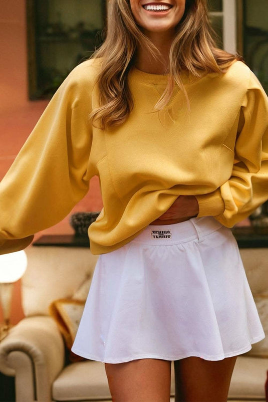 Woman wearing citrus yellow pleated balloon sleeve pullover sweatshirt with white skirt indoors