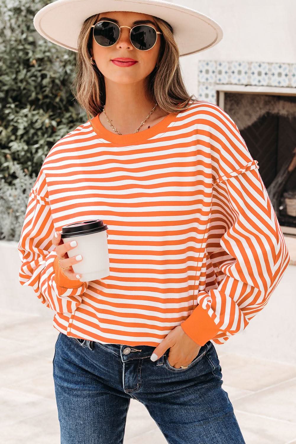 Woman wearing orange stripe exposed seam drop shoulder casual pullover top with sunglasses and hat holding a coffee cup